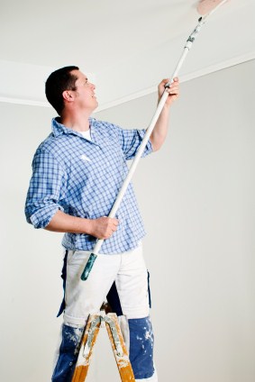 Painter on step ladder painting ceiling with a roller on a pole.